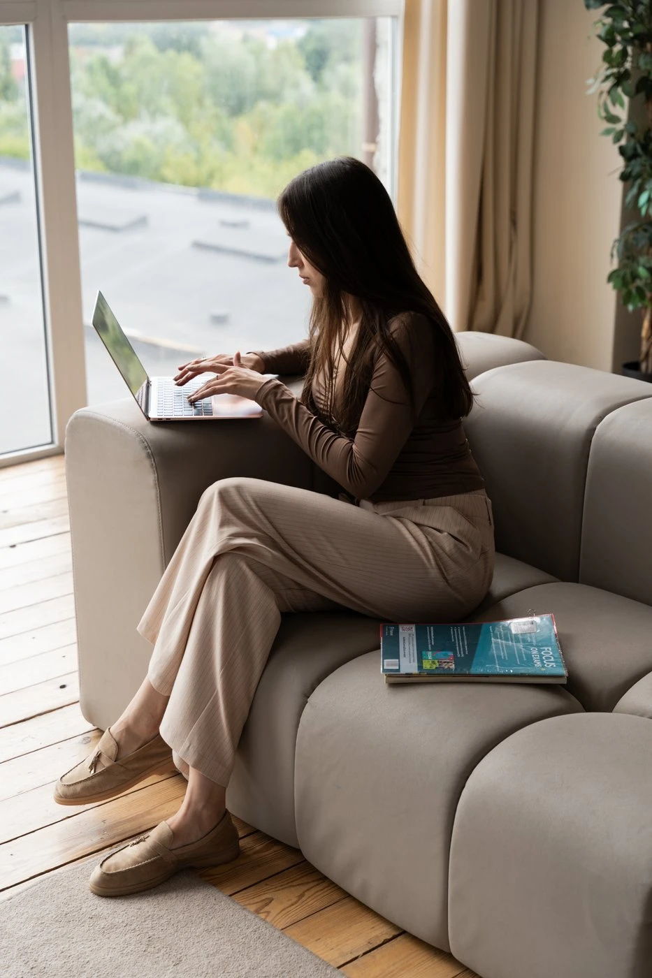 Woman sitting on a sofa with a laptop, typing, next to large windows. A magazine rests on the sofa beside her.